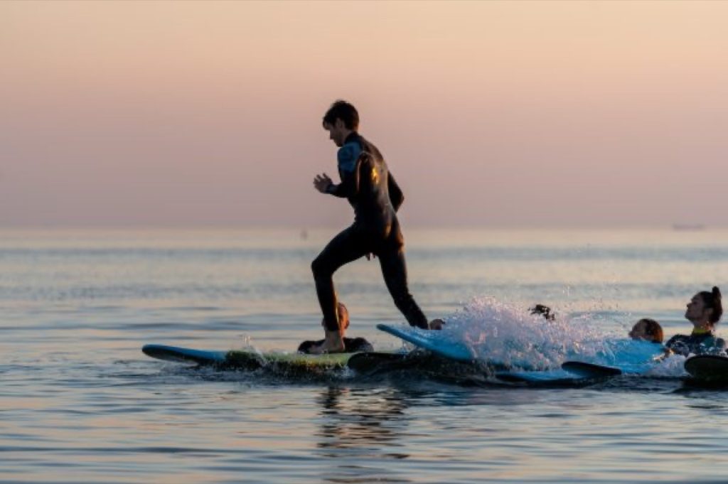 Banglentininkas ant banglentės saulėlydžio metu, „Surf Camp“